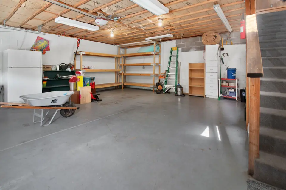 Garage with a plain gray concrete floor, wood storage shelves, a refrigerator, a ladder and a wheelbarrow