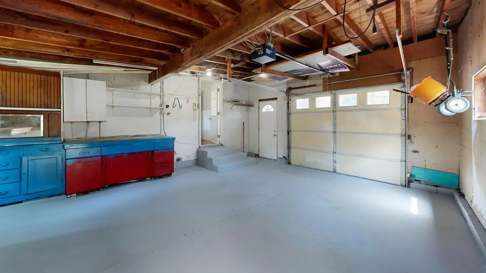 Older one-car garage with open wood joists, a sectional door, blue and red cabinets and a flat light-gray painted concrete floor with a hairline crack